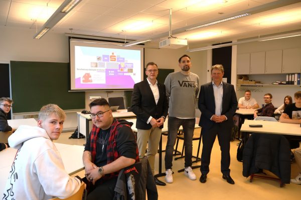 zu sehen ist Stehend sind v.l.n.r. Herr Dr. Sören Abendroth (Vorsitzender der Sparkasse Mittelholstein), Carsten Janz (Journalist und Dozent) und Herr Marc-Olaf Begemann (Schulleiter des BBZ am NOK) umgeben von den Teilnehmern des Workshops. (Foto: Stephan Doemeland)