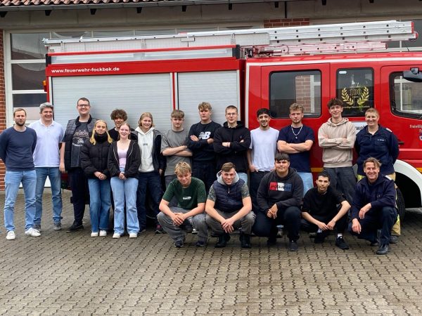 TM 1 Lehrgang Berufsfachschule Technik 2025 (Foto (Andreas Gädigk): Von links: Berufsschullehrer Nico Greve und Ralph Stangenberg mit den erfolgreichen Schülerinnen und Schülern der Berufsfachschule I Technik sowie Sandra Gädigk (FF Fockbek) und Amtswehrführer Dr. Jan Traulsen (rechts kniend))
