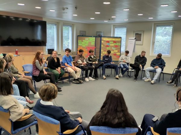 Eine Gruppe von deutschen und französische Schülern sitzen in einem Stuhlkreis in einem Konferenzraum. Zwischen den Schülern sitzt der Moderatorin, die den Austausch moderiert.