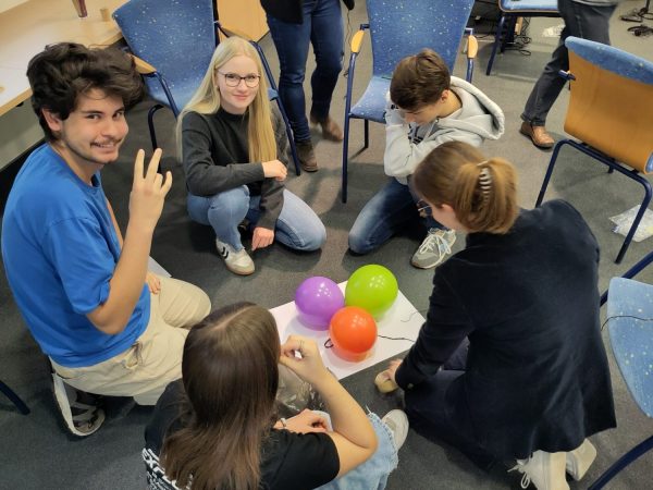 Fünf Schülerinnen und Schüler sitzen und knien auf dem Boden in einem Konferenzraum um ein Stück Papier auf dem 3 farbige Luftballons liegen.
