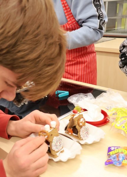 Ein Schüler dekoriert ein kleines aus Spekulatiuskeksen dreiecksförmiges Lebkuchenhaus. Daneben steht bereit ein weiteres fertiges Haus.