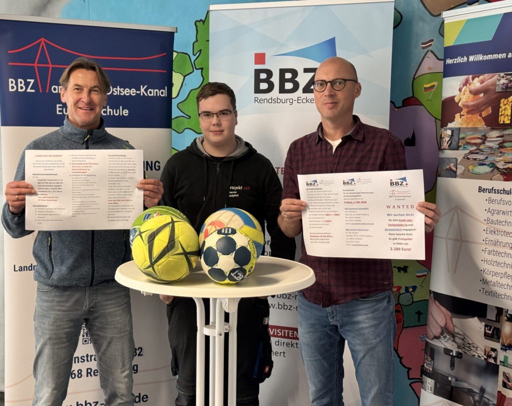 links steht Jan Traulsen und hält einen ausgefalteten Flyer vor sich, rechts daneben steht der Auszubildende Jean-Etienne Westphal, der hinter einem weißen Stehtisch mit 4 Fußbällen steht. Rechts daneben steht Arne Fiebelkorn der die andere Seite eines Flyers vor sich hält.