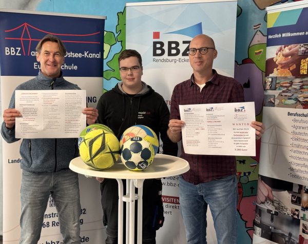 links steht Jan Traulsen und hält einen ausgefalteten Flyer vor sich, rechts daneben steht der Auszubildende Jean-Etienne Westphal, der hinter einem weißen Stehtisch mit 4 Fußbällen steht. Rechts daneben steht Arne Fiebelkorn der die andere Seite eines Flyers vor sich hält.