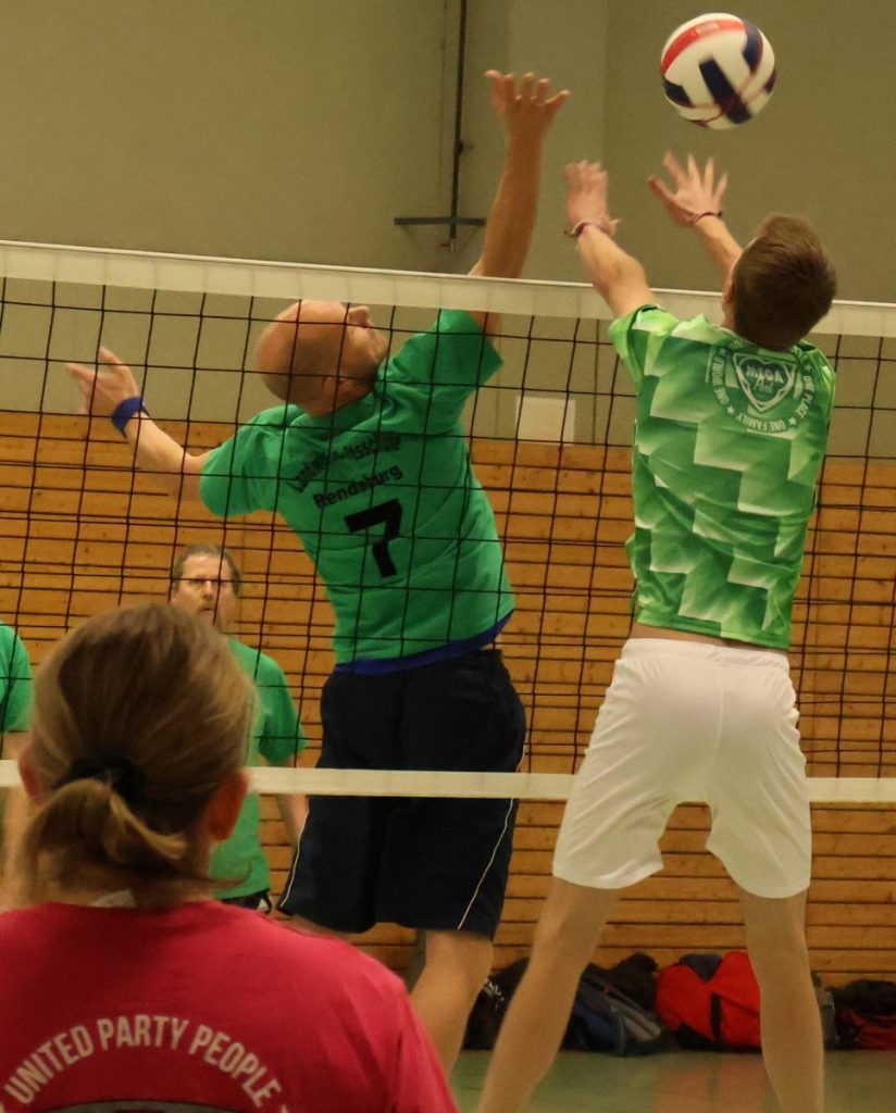 Der Spieler hinter dem Netz steht mit dem Rücken zum Netz und spielt den Ball mit einer Hand über das Netz. Der Spieler auf dieser Seite des Netzes ist ebenfalls hochgesprungen, um den Ball zu blocken. Im Vordergrund steht eine Spielerin und beobachet die Szene.