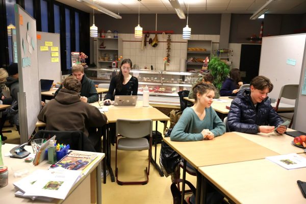 Sieben Schüler sitzen in Kleingruppen an an jeweils an vier zusammengestellten Tischen und arbeten in Kleingruppen. Im Vordergrund sitzen zwei Schüler und arbeiten. Schräg links dahinter sitzen 3 Schüler zusammen und rechts dahinter sitzen zwei Schüler zusammen.