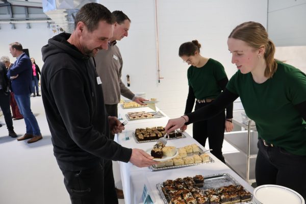 Zwei Kollegen des BBZ stehen am Kuchenbufett. Ihnen gegenüber stehen zwei junge Frauen aus dem Bereich Hauswirtschaft im ländlichen Raum mit grünen Oberteilen. Die vordere legt einer Lehrkraft des BBZ gerade ein Kuchstück auf den Teller.