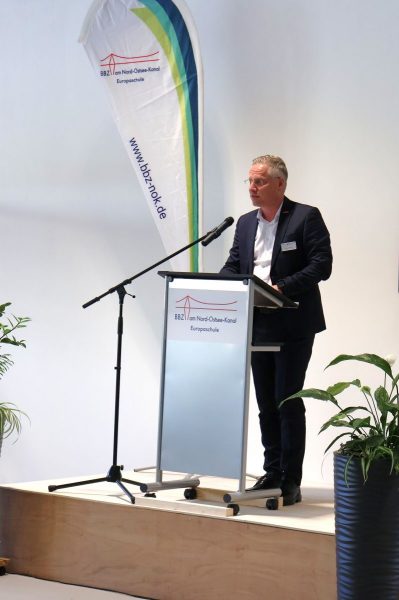 Herr Björn Geertz steht auf einem Podest in einem dunklen Anzug und weißem Hemd. Vor ihm ist ein Rednerpult mit dem Logo des BBZ-NOK. Links neben ihm steht eine Beachflag ebenfalls mit dem Logo des BBZ-NOK.