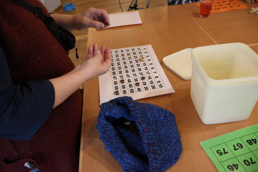 Auf einem bruanen Holztisch liegt ein Bingo Zahlenfeld 1-70 mit einigen Steinen darauf. Im Vordergrund ist ein blauer Beutel zu sehen. Daneben steht ein hohe quadratische weiße Kunstoffbox. Es sind die Hände von einer Person zu sehen, die einen Nummernstein in der linken Hand hält.