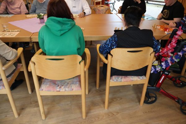 EIne Schülerin mit grünem Hoddie sitzt am Tisch mit mehreren Personen. Neben ihr Sitz eine Seniorin. Neben der Seniorin steht einen mit Blumenkette geschmückter Rollator.