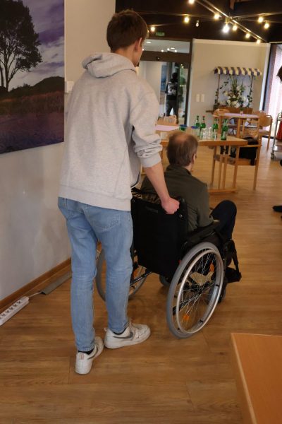 Ein Schüler mit wißem Hoddie und blauer Jeans, schiebt einen älteren Herren in seinem Rollstuhl Richtung einem Durchgang.