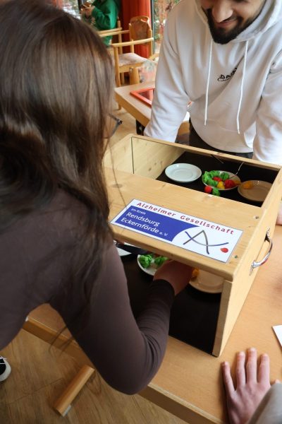 Eine braunhaarige Schülerin mir einem braunen Pullover steht gebückt an einem Holzkasten mit Spiegel an der Rückwand. Es ist Geschirr im Kasten zu erkennen. Der direkte Blick auf das Geschirr, wir durch ein Brett versperrt, auf dem ein Aufkleber der Altzheimer Gesellschaft zu sehen ist.