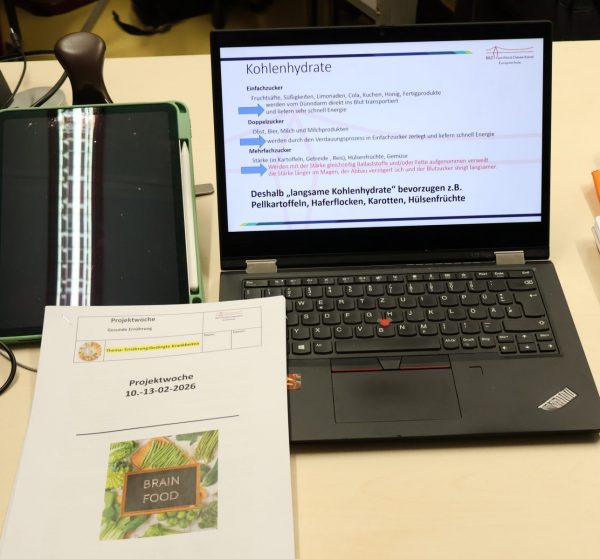 Ein Tisch mit einem Laptop, auf dem Informationen über Kohlenhydrate zu sehen ist. Daneben ein Tablet. Halt auf der Tastatur liegt ein Zettel der Projektwoche "Gesunde Ernährung" Thema "Ernährungsbedingte Krankheiten"
