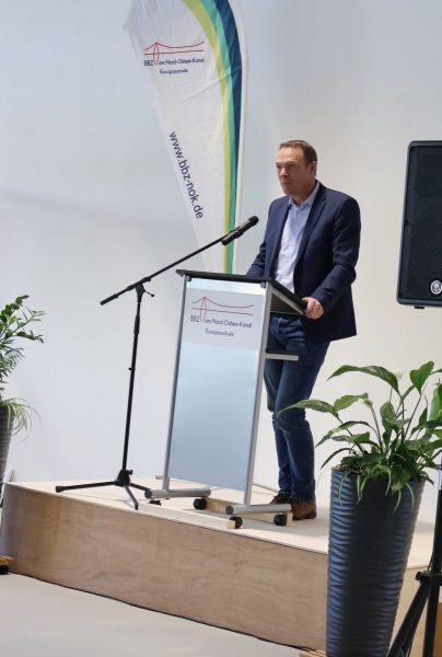 Kreisrat Ingo Sander steht auf einem Podest in einem dunklen Anzug und weißem Hemd. Vor ihm ist ein Rednerpult mit dem Logo des BBZ-NOK. Links neben ihm steht eine Beachflag ebenfalls mit dem Logo des BBZ-NOK.