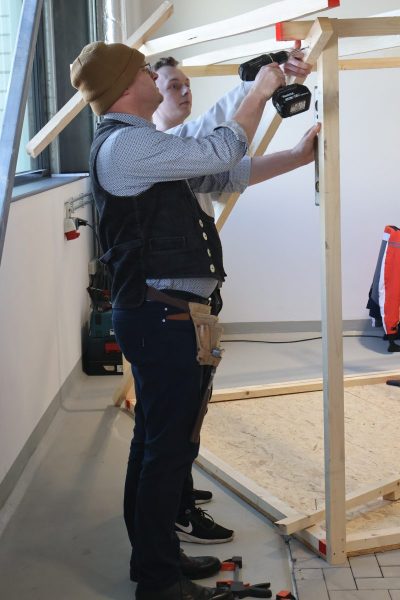 Zwei männlichen Kollegen des BBZ-NOK stehen neben einer Holzkonstruktion in der neuen Bauwerkstatt. Der vordere Bohr mit einer Akkubohrmaschine ein Loch in eine Holzlatte, die von unten schräg nach oben führt. Wärend der ander Kollege die Holzlatte hält.