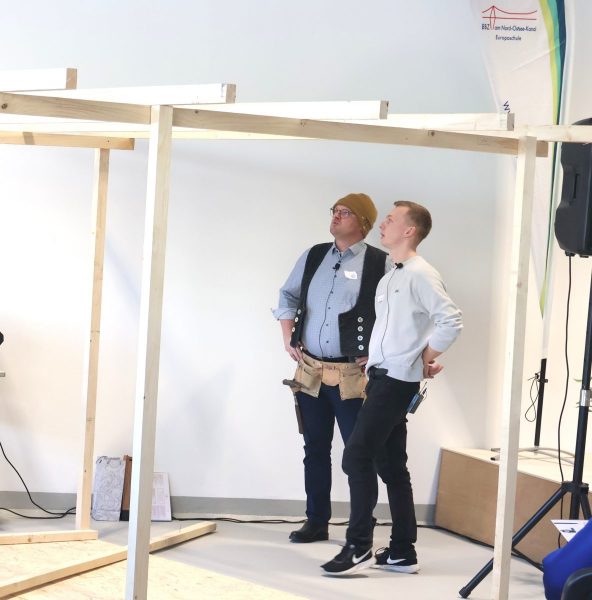 Zwei männliche Kollen des BBZ-NOK stehen in einem hellen weißen Raum unter einer Holzkonstruktion in den Räumen der neuen Bauwerkstatt.