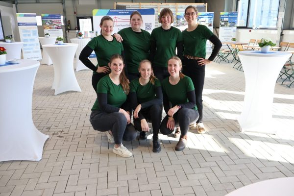 Sieben junge angehende Betriebsleiterinnen der Hauswirtschaft im ländliche Raum. Die einem Raum mit vielen weißen Stehtischen als Team posieren. Alle tragen ein grünes Oberteil. Vier von Ihnen stehen in der hinteren Reihe, wärend 3 davor in der Hocke sind und in die Kamera lächeln.