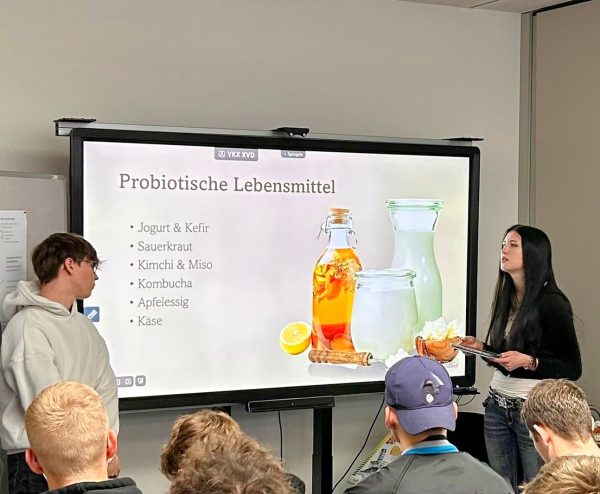 Zwei Schüler stehen rechts und links von einem Smartboard. Auf dem Bildschirm ist unter der Überschrift Probiotische Lebensmittel eine Auflistung zu sehen. Im Vordergrund sind sitzen vier Schüler und hören dem der Präsentation zu.