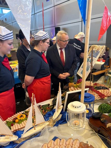 Jurymitglied in Anzug und roter Krawatte begutachtet mit zwei Personen in schwarzem Hemd und roter Schürze ein maritim dekoriertes Buffet mit Segelbootmodellen, Laterne und verschiedenen kalten Speisen.