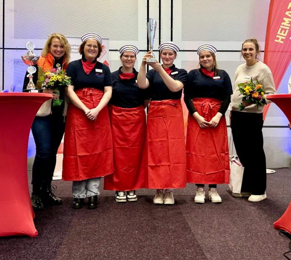 Eine Gruppe von fünf jungen Frauen steht nebeneinander und lacht. Die vierte von links hält einen silbernen Pokal über ihren Kopf. Alle bis auf die beiden äußeren Frauen tragen identische dunkelblaue Hemden, rote Schürzen und gestreifte Mützen. Die Frau ganz links im Bild, wahrscheinlich eine Betreuerin, trägt ein orangefarbenes Oberteil, eine dunkle Jeans und Stiefel und hält einen Blumenstrauß und einen kleinen Pokal. Die Frau ganz rechts, ebenfalls eine Betreuerin, trägt einen hellen Pullover, eine schwarze Hose und hält ebenfalls einen Blumenstrauß. Alle Frauen sind kaukasischer Abstammung und im jungen Erwachsenenalter. Sie stehen auf einem dunkelbraunen Teppich in einem Innenraum, der von leuchtend roten Tischdecken an den Seiten und einer Fahne mit der Aufschrift "HEIMAT" im Hintergrund erhellt wird. Die Beleuchtung ist hell und gleichmäßig, was eine fröhliche und feierliche Atmosphäre schafft. Die Perspektive ist auf Augenhöhe, und die Aufnahme ist zentriert auf die Gruppe, wobei die Frauen den Großteil des Bildes ausfüllen.