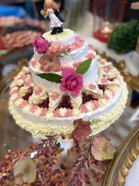 Eine mehrstöckige Torte, die anscheinend aus Käse besteht, ist reichhaltig mit weißer und rosa Creme verziert und mit frischen, leuchtend pinken Rosen geschmückt. Auf der Spitze thront eine klassische Hochzeitspaar-Figur, während die Basis von getrockneten Blüten und Dekorationen umgeben ist.