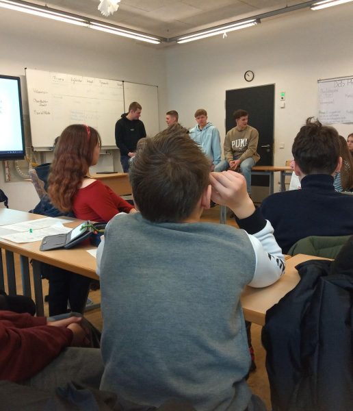 Mehrere Jugendliche sitzen und stehen in einem Klassenraum. Im Vordergrund ist der Hinterkopf einer Person mit kurzen Haaren zu sehen, die an einem Tisch sitzt. Im Hintergrund stehen vier Jugendliche auf Tischen, während andere auf Stühlen sitzen und zuhören. An den Wänden hängen weiße Tafeln mit handschriftlichen Notizen und ein Plakat mit dem Titel 'Das AIDA'. Die Szene zeigt eine Präsentation zum Thema Landwirtschaft in einer 7. und 8. Klasse.