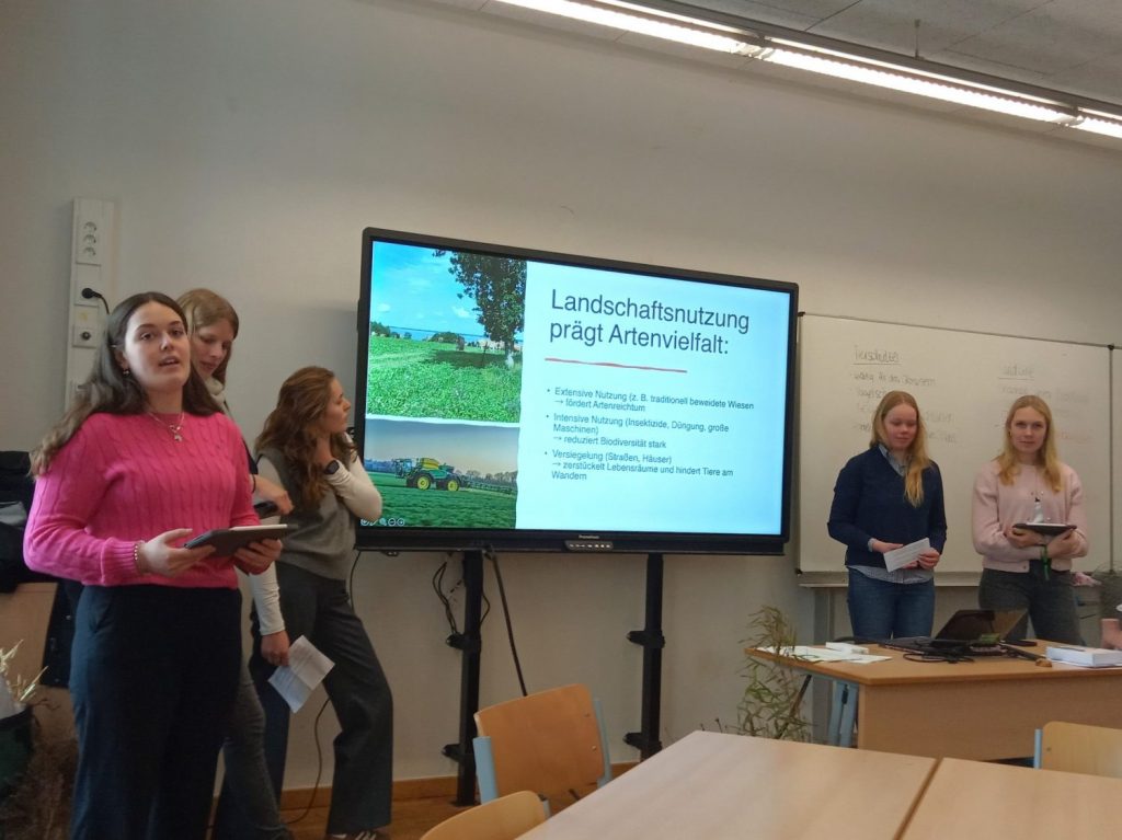 Vier junge Frauen stehen in einem Klassenraum und halten Präsentationskarten oder Tablets in den Händen. Im Hintergrund befindet sich ein großer Bildschirm, auf dem eine Präsentation mit dem Titel 'Landschaftsnutzung prägt Artenvielfalt:' gezeigt wird. Der Bildschirm zeigt außerdem zwei Farbfotos von landwirtschaftlichen Flächen und einen Traktor. Die Tafel rechts im Bild ist teilweise beschrieben. Die Szene vermittelt den Eindruck einer Gruppenpräsentation im Rahmen einer Schulveranstaltung. Der Kontext 'Maritimes Buffet' ist nicht sichtbar, daher liegt der Fokus auf der Präsentationssituation.