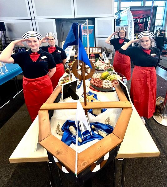 Buffet in Schiffsform mit maritimen Dekorationen und vier Personen in schwarzen T-Shirts, roten Schürzen und roten Halstüchern, die salutieren