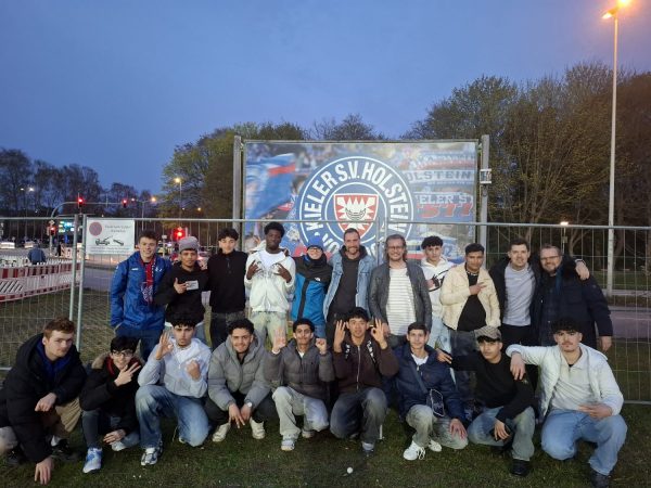Vor dem Holsteinstadion in Kiel steht eine Gruppe von Lehrern und Schülern der Fußball AG. Im Hintergrund ist ein großes Schild mit dem Vereinslogo des Kieler SV Holstein und dem Schriftzug 'Holstein Kiel' zu sehen. Die Personen sind unterschiedlich gekleidet, einige tragen Sportkleidung, andere Freizeitkleidung. Sie stehen und hocken auf dem Rasen, viele lächeln und zeigen Handzeichen. Die Szene spielt am Abend, das Licht ist bläulich und die Umgebung zeigt Bäume sowie Straßen mit Ampeln.