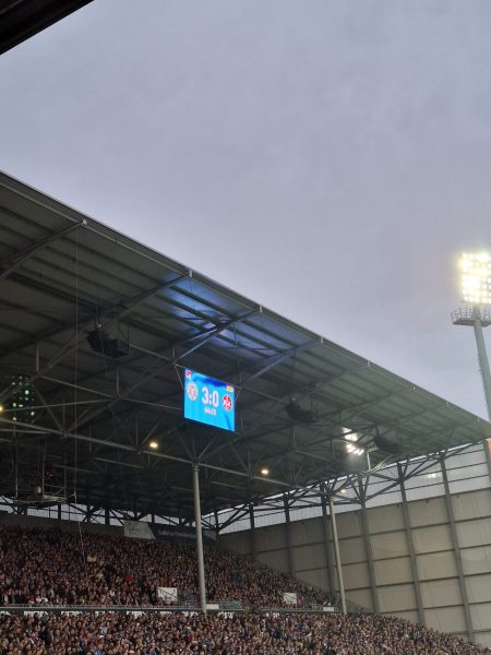 Im Holsteinstadion zeigt eine elektronische Anzeigetafel den Spielstand 3:0 für Holstein Kiel gegen 1. FC Kaiserslautern. Die Tribüne ist dicht besetzt, das Stadiondach und ein Flutlichtmast sind sichtbar. Der Himmel ist grau und bewölkt.
