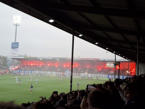 Die Tribüne des Holsteinstadions ist dicht gefüllt, zahlreiche Fans halten rote Bengalos in die Höhe und sorgen für eine intensive Atmosphäre. Im Vordergrund stehen Zuschauer unter dem überdachten Bereich, viele filmen oder fotografieren mit ihren Smartphones. Auf dem Spielfeld laufen Spieler von Holstein Kiel und dem gegnerischen Team, während im Hintergrund ein großes Banner mit gut lesbarer Schrift quer über die Tribüne gespannt ist. Die Flutlichtanlage leuchtet hell, und der Himmel wirkt grau und diesig.