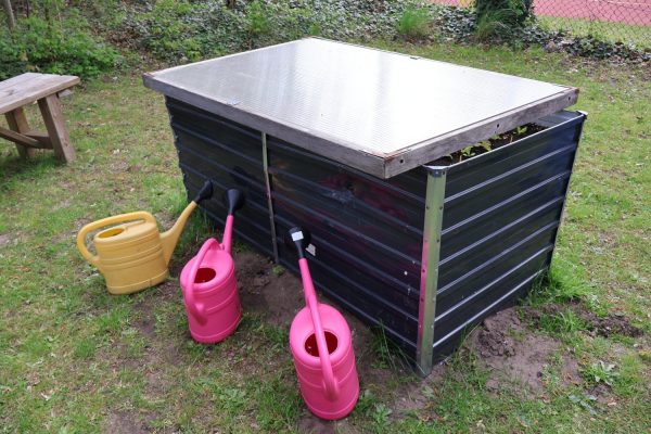 Rechteckiges Hochbeet mit dunklen, waagerechten Seitenwänden aus Metall oder Kunststoff und einer silberfarbenen Abdeckung. Drei Gießkannen in Gelb und Pink stehen davor auf dem Rasen. Im Hintergrund sind eine Holzbank, Rasenfläche und eine Hecke zu sehen.