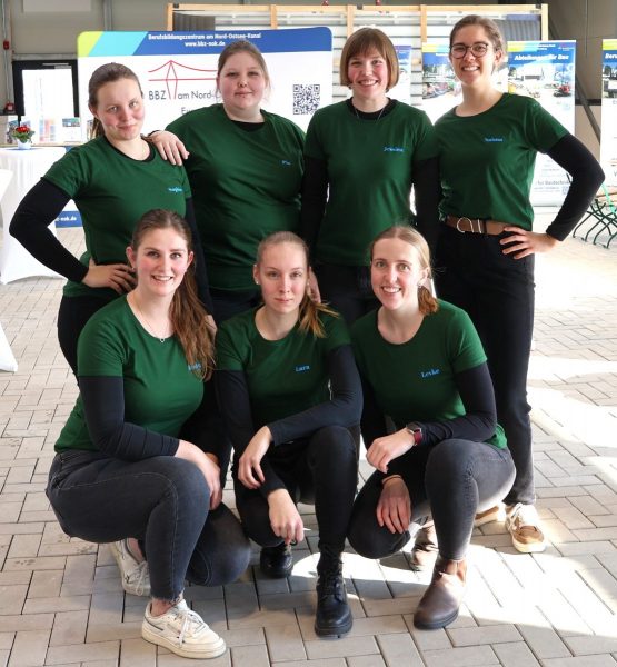 Sieben angehende Betriebsleiterinnen der Fachschule für Hauswirtschaft stehen und knien in zwei Reihen in einem Innenraum mit hellen Bodenfliesen. Alle tragen dunkelgrüne T-Shirts mit Namensaufdruck und schwarze Hosen. Im Hintergrund sind Informationsstände mit Plakaten, Tischen und Stühlen zu sehen. Die Frauen blicken in Richtung Kamera, ihre Gesichter sind unkenntlich gemacht.
