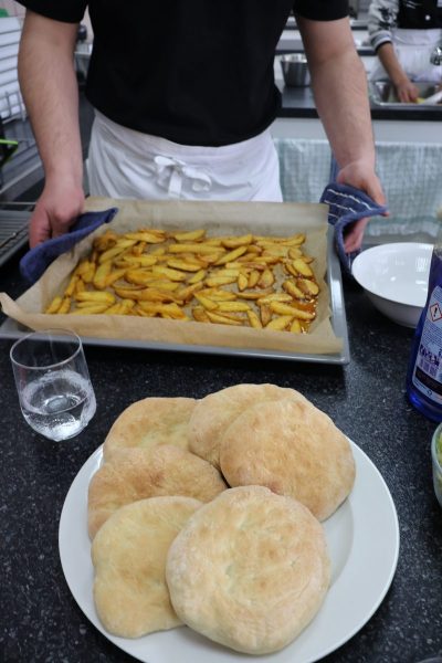 Im Vordergrund liegt ein weißer Teller mit mehreren runden, flachen Broten auf einer dunklen Arbeitsfläche. Dahinter hält eine Person, die eine weiße Schürze trägt, ein Backblech mit frisch gebackenen Kartoffelspalten, die auf Backpapier liegen. Ein Glas Wasser und eine geöffnete Flasche stehen daneben. Die Szene zeigt eine praktische Übung der angehenden Betriebsleiterinnen der Fachschule für Hauswirtschaft.