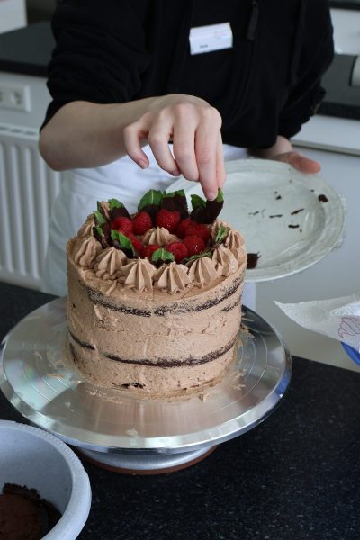 Eine angehende Betriebsleiterin der Fachschule für Hauswirtschaft steht an einer Arbeitsfläche und dekoriert eine mehrschichtige Torte mit Schokoladencreme. Die Torte ist mit Sahnetupfen und frischen Himbeeren sowie grünen Blättern verziert. Die Person trägt dunkle Oberbekleidung und eine weiße Schürze. Im Hintergrund sind Küchenutensilien und ein weißer Heizkörper zu sehen.