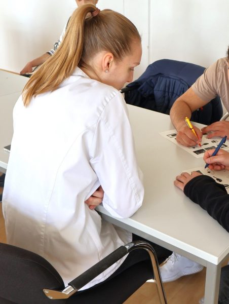 Eine angehende Betriebsleiterin der Fachschule für Hauswirtschaft sitzt an einem Tisch. Sie trägt einen weißen Kittel und hat die Haare zu einem Pferdeschwanz gebunden. Vor ihr liegt ein Blatt Papier, auf das mehrere Personen mit Stiften zeigen oder darauf schreiben. Die Szene spielt in einem hellen Raum mit weißen Wänden und Holzboden. Die Atmosphäre wirkt sachlich und konzentriert.