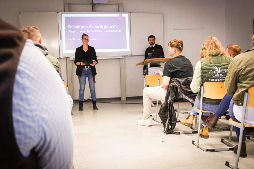 Im Vordergrund sitzen mehrere Personen auf gelben Stühlen und blicken nach vorn. Im Zentrum steht eine Frau, laut Kontext Rebecca Meurer, die vor einer Projektionsfläche spricht und ein Tablet oder einen Block in den Händen hält. Rechts neben ihr steht ein Mann an einem Stehpult. Auf der Leinwand ist der Titel der Veranstaltung zu lesen: 'Fachforum Klima & Umwelt – Wege zu einer klimastabilen Landwirtschaft'. Die Atmosphäre wirkt sachlich und konzentriert, typische Merkmale eines Vortrags oder einer Diskussionsrunde in einem Klassenraum.