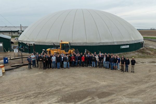 Vor einem großen, halbrunden Gebäude mit weißer Kuppel und grünem Sockel, das als Biogasanlage der Eilers Farm identifiziert wird, steht eine große Gruppe von Menschen in mehreren Reihen. Im Hintergrund ist ein gelber Radlader zu sehen. Der Boden besteht aus festem, hellbraunem Kies. Die Szene spielt im Freien bei bedecktem Himmel.