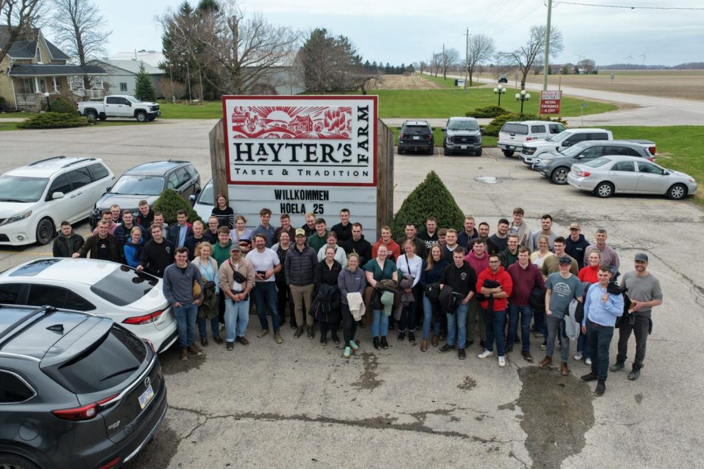 Vor dem großen Schild mit der Aufschrift 'HAYTER'S FARM TASTE & TRADITION' und dem darunter angebrachten Begrüßungsschild mit dem Text 'WILLKOMMEN HOELA 25' steht eine große Gruppe von Menschen. Die Gruppe besteht aus Jugendlichen und Erwachsenen, die eng beieinander stehen und in die Kamera blicken. Im Hintergrund sind mehrere geparkte Autos, ein Gebäude mit Veranda und eine weitläufige ländliche Umgebung mit Feldern und Bäumen zu sehen. Die Szene spielt tagsüber bei bewölktem Himmel.