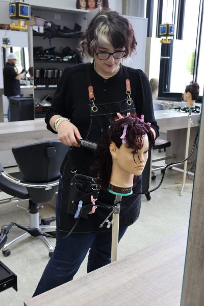 Eine angehende Friseurin steht in einem modernen Friseursalon und arbeitet an einem Übungskopf, der auf einem Stativ befestigt ist. Sie trägt eine dunkle Latzschürze mit mehreren Friseurwerkzeugen, darunter Klammern und eine Bürste. Die Haare des Übungskopfes sind mit mehreren farbigen Klammern abgeteilt. Im Hintergrund sind Spiegel, Regale mit Produkten und weitere Übungsköpfe zu sehen. Die Szene zeigt eine praktische Übung im Rahmen des Lernfeldes „Komplexe Friseurdienstleistungen durchführen“.