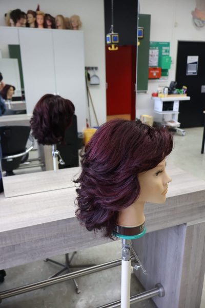 Im Vordergrund steht ein Friseurübungskopf mit dunkelbraunem, leicht violett schimmerndem, schulterlangem Haar, das in weichen Stufen geschnitten und nach außen geföhnt wurde. Die Frisur wirkt voluminös und gepflegt. Im Hintergrund sind Spiegel, Friseurstühle, ein Regal mit Friseurprodukten sowie mehrere Übungsköpfe auf einem Schrank zu sehen. Die Umgebung erinnert an einen modernen Friseursalon, passend zum Kontext der Ausbildung im Bereich komplexer Friseurdienstleistungen.
