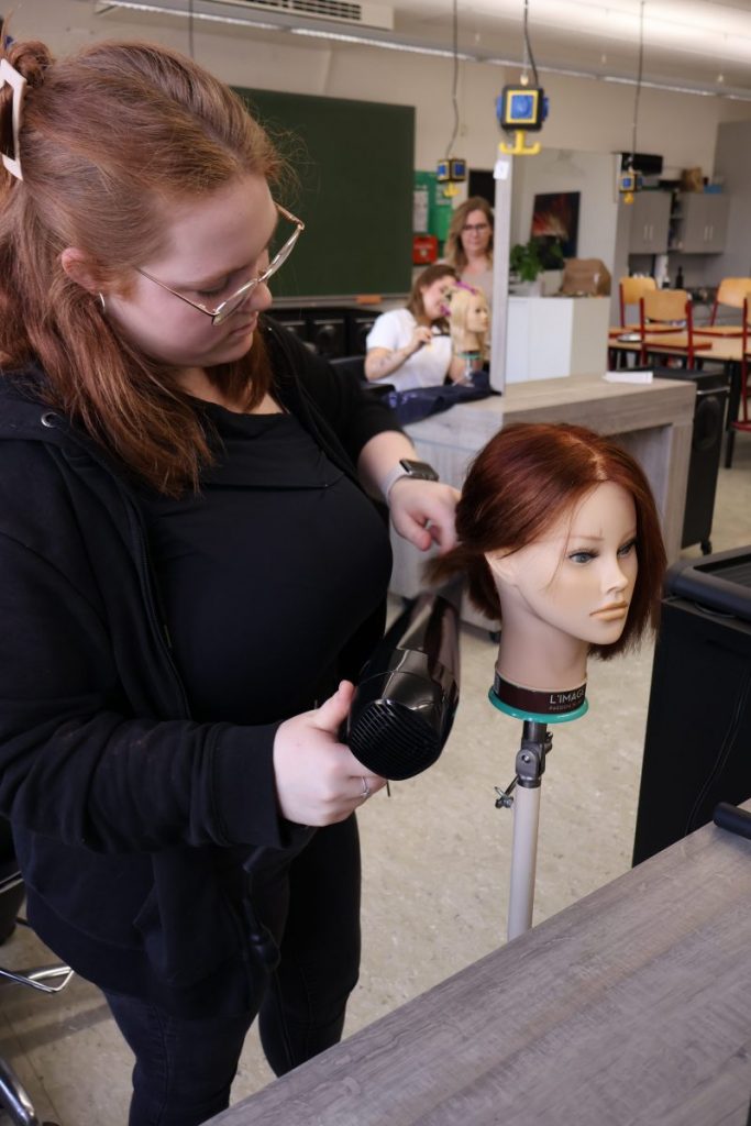 Eine angehende Friseurin hält einen Föhn in der rechten Hand und arbeitet an einem Übungskopf mit braunem, kinnlangem Haar. Sie trägt schwarze Kleidung und eine Brille. Im Hintergrund befinden sich weitere Personen, die ebenfalls an Übungsköpfen arbeiten. Die Szene spielt in einem modern ausgestatteten Klassenraum mit mehreren Tischen, Stühlen und Friseurbedarf. Die Beleuchtung ist hell und gleichmäßig.