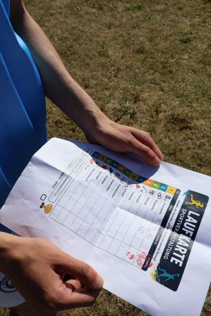 Eine Person hält eine Laufkarte in den Händen, die für das Sport- und Spielevent der Berufsvorbereitungsklassen am BBZ am NOK genutzt wird. Die Laufkarte ist auf weißem Papier gedruckt und zeigt eine Tabelle mit verschiedenen Stationen, die jeweils durch farbige Symbole und Zahlen gekennzeichnet sind. Einige Felder sind handschriftlich ausgefüllt oder abgestempelt. Im oberen Bereich der Karte steht in großen, gut lesbaren Buchstaben 'LAUFKARTE' sowie 'SPORTFEST WERTUNG'. Die Person trägt ein ärmelloses, hellblaues Oberteil. Der Hintergrund besteht aus trockenem, braunem Rasen, was auf eine Outdoor-Veranstaltung bei sonnigem Wetter hindeutet.
