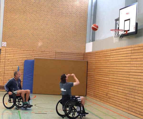Jacob Zuleger sitzt in einem Rollstuhl und führt einen Korbleger mit einem Basketball in einer Sporthalle aus. Eine weitere Person im Rollstuhl beobachtet die Szene. Die Halle hat eine Holzverkleidung an den Wänden, eine Basketballanlage mit weißem Brett und orangem Ring sowie farbige Linien auf dem Hallenboden. Im Hintergrund ist eine blaue Matte an der Wand zu sehen.