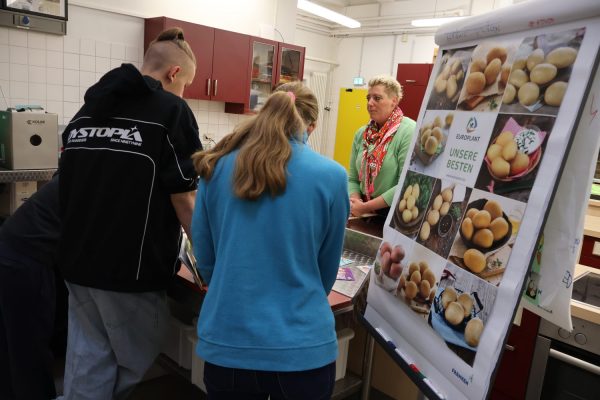 Im Vordergrund steht eine große Infotafel mit Fotos verschiedener Kartoffelsorten. Die Bilder zeigen unterschiedliche Formen und Farben der Kartoffeln, jeweils mit kurzen Beschriftungen. Mehrere Personen stehen um einen Küchentisch und beschäftigen sich mit dem Thema Kartoffeln. Im Hintergrund sind rote Küchenschränke, eine gelbe Tür und eine Person mit einem bunten Schal zu sehen. Die Szene spielt in einer Schulküche, die für den Fachpraxisunterricht genutzt wird.