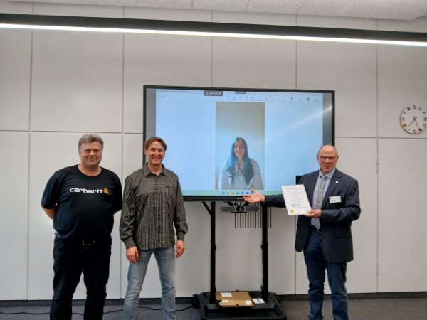 Drei Personen stehen in einem modernen, hellen Raum vor einem großen Bildschirm. Auf dem Bildschirm ist eine Frau zu sehen, die freundlich in die Kamera blickt. Die Person ganz rechts hält eine Urkunde in der Hand und wendet sich den anderen zu. Die mittlere Person trägt ein kariertes Hemd, die linke ein dunkles T-Shirt mit einem gut lesbaren Markenschriftzug. Im Hintergrund sind eine weiße Wand mit vertikalen Linien und eine analoge Wanduhr zu erkennen. Die Szene vermittelt den Moment einer Auszeichnung, bei der Simone Bock als Landessiegerin der Elektroinnung Rendsburg geehrt wird. Sönke Lucht, Dr. Jan Traulsen und Michael Petersen sind an der Ehrung beteiligt.