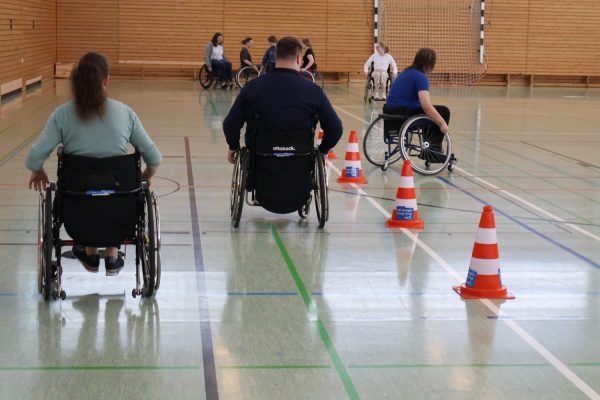Mehrere Jugendliche in Sportrollstühlen bewegen sich in einer Sporthalle durch einen Slalomparcours aus orange-weißen Pylonen. Die Halle hat eine Holzverkleidung an den Wänden und einen hellen Boden mit farbigen Linien. Im Hintergrund sind weitere Teilnehmende in Rollstühlen zu sehen. Die Szene zeigt eine inklusive Sportveranstaltung im Rahmen des Projekts „Rollstuhlsport macht Schule“ am BBZ am NOK, organisiert in Kooperation mit dem Deutschen Rollstuhl-Sportverband e. V. und der Unfallkasse Nord. Die Jugendlichen tragen sportliche Alltagskleidung.