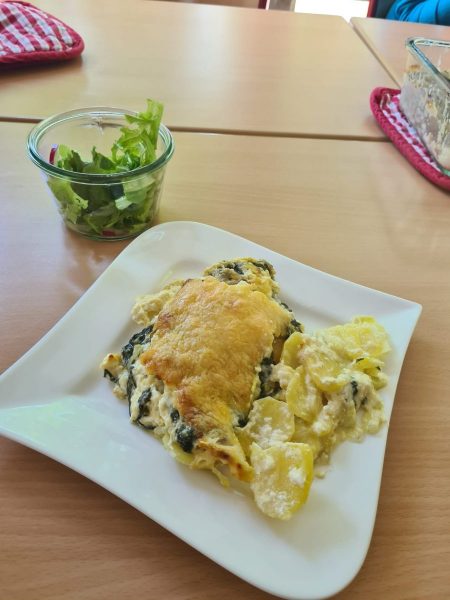 Auf einem hellen Holztisch steht ein weißer, eckig geschwungener Teller mit einer Portion Spinat-Kartoffelauflauf, der mit goldgelb gebackenem Schafskäse überbacken ist. Die Schichten aus Kartoffelscheiben und Spinat sind deutlich zu erkennen. Daneben steht ein kleines Glas mit frischem grünen Salat. Im Hintergrund liegen rote Topflappen und eine leere Auflaufform. Das Gericht wurde im Rahmen des Fachpraxisunterrichts der Ausbildungsvorbereitungsklassen AVSH-i a und b mit Gemüse aus dem Schulgarten zubereitet.