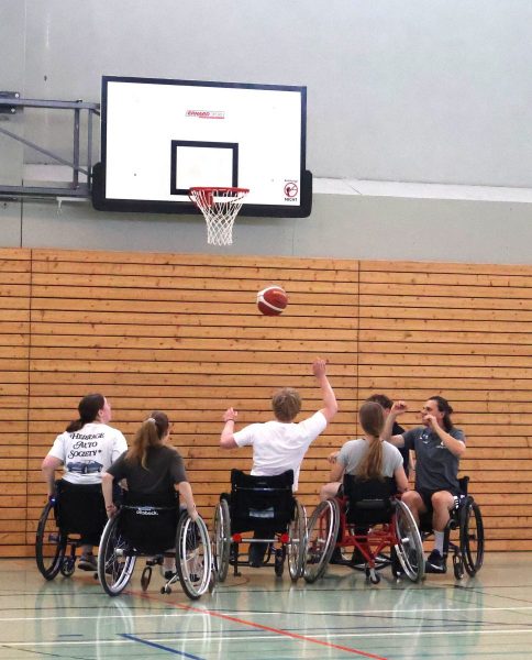 Fünf Jugendliche in Sportrollstühlen befinden sich in einer Turnhalle vor einem Basketballkorb. Sie tragen Sportkleidung, und eine Person wirft einen Basketball in Richtung Korb. Die Gruppe ist in einer Formation angeordnet, die auf eine taktische Spielsituation im Rollstuhlbasketball schließen lässt. Im Hintergrund sind eine Holzprallwand und ein Basketballbrett mit Netz zu sehen. Die Szene dokumentiert eine inklusive Sporteinheit im Rahmen des Projekts „Rollstuhlsport macht Schule“ am BBZ am NOK, organisiert von Jacob Zuleger in Kooperation mit dem Deutschen Rollstuhl-Sportverband und der Unfallkasse Nord.