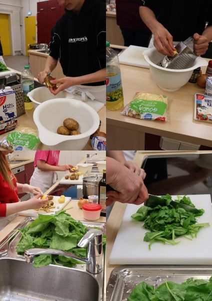 Mehrere Jugendliche bereiten in einer Schulküche frisches Gemüse zu. Auf der Arbeitsfläche liegen Kartoffeln, ein großer weißer Kunststofftopf, eine Reibe, ein Schneidebrett und verschiedene Zutaten wie Öl, Sahne und Kräuter. Im Vordergrund wird Blattgemüse auf einem weißen Schneidebrett geschnitten. Die Szene zeigt praktische Küchenarbeit im Rahmen des Fachpraxisunterrichts der Ausbildungsvorbereitungsklassen AVSH-i a und b in Kooperation mit der Schule Hochfeld. Die Zutaten stammen aus dem Schulgarten.