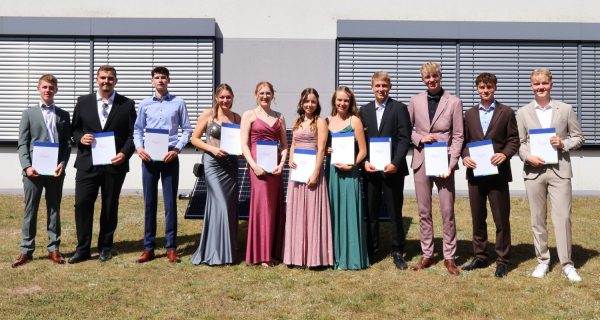 Eine Gruppe junger Menschen steht nebeneinander auf einer Rasenfläche vor einem modernen Gebäude mit geschlossenen Jalousien. Alle tragen festliche Kleidung, darunter Anzüge und lange Abendkleider. Jede Person hält ein Dokument mit blauem Rand in den Händen, das auf eine Abschlussfeier hindeutet. Die Szene wirkt feierlich und zeigt den Moment der Zeugnisübergabe für den Abschlussjahrgang des Beruflichen Gymnasiums Erneuerbare Energien am BBZ am NOK.